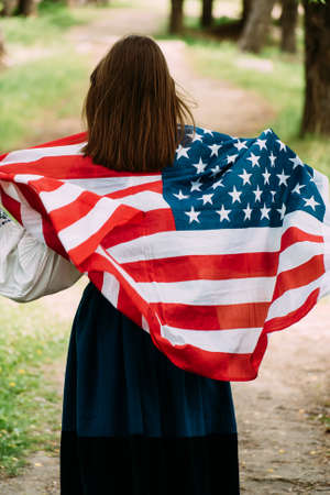 Woman posing with USA national flag standing outdoors in summer park. Positive girl celebrating United States independence day.の写真素材