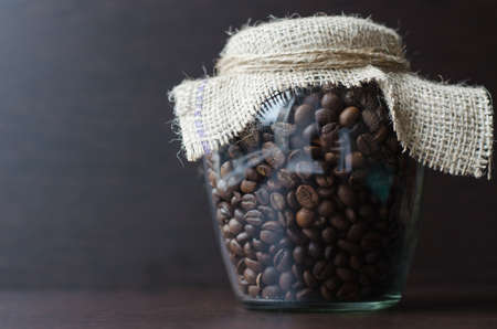 Large transparent jar with coffee beans closeup. Bank with coffee on a wooden table. Place for your...