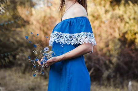 A young woman in a blue dress holds in her hands a bouquet of wildflowers. A girl is standing in the...