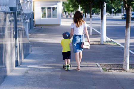 woman with a child are walking along the street, rear view. The child rides on a green scooter rear viewの写真素材