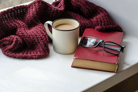 white cup with coffee, book, glasses and burgundy knitted scarf by the window.