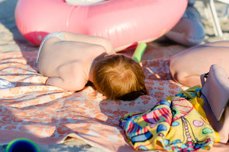A small child with red hair sleeps on the beach. The child is curled up in a ball and sleeps under the rays of the sun.の写真素材