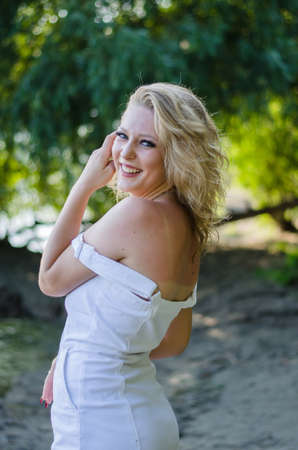 Beautiful woman looking at the camera and smiling. Blonde in white dress in natureの写真素材