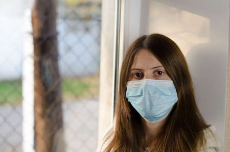A young woman in a medical mask sits by the window. Self-isolation concept. Prevention covid 19
