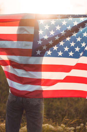 Rear view of a young woman holding the US national flag in a field at sunset. Independence Day of...