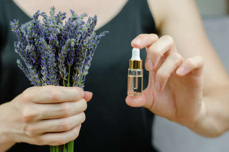 A bottle with lavender oil in female hands. A woman in black holds a transparent bottle of essential...