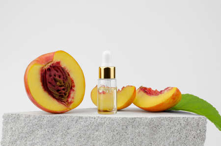 A transparent bottle with a dropper with peach oil and fresh peaches on a gray podium. Natural skin and hair care. Natural cosmeticの写真素材