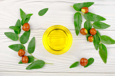Jojoba oil in a bowl, a sprig of jojoba and fruits on a white wooden tableの写真素材