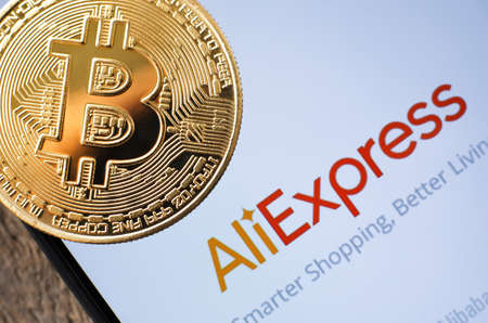 A gold Bitcoin coin lies on a smartphone with the emblem of the Aliexpress corporation - Ukraine,...