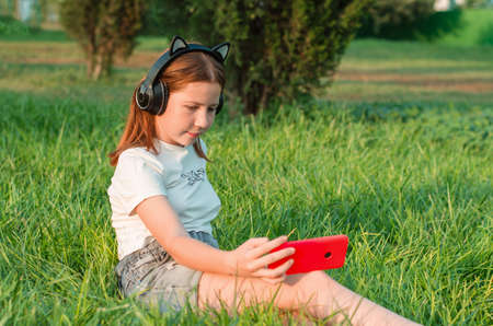 A teenage girl sits in a park on the grass with a smartphone in her hands. 12 year old girl listening to music in headphones sitting in the park on the grassの写真素材