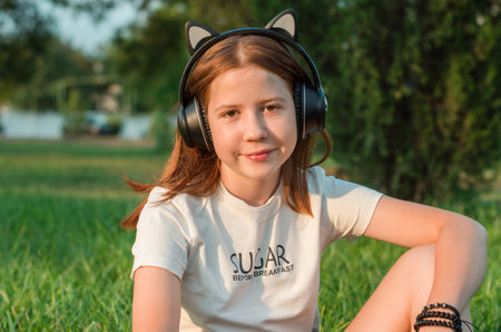 Portrait of a beautiful red-haired teenage girl in headphones in nature. 12 year old girl listens to music while sitting on the grass in the park. happy childhood.の写真素材