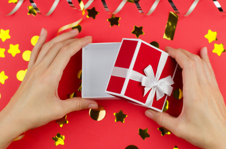 Open gift box in female hands on a red festive background, close-up. Woman opening a small gift boxの写真素材