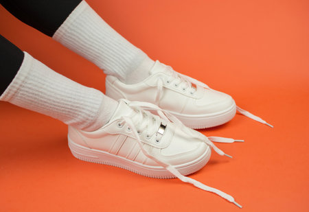 Women's legs in stylish white sneakers with untied laces. Sneakers on legs on orange backgroundの写真素材