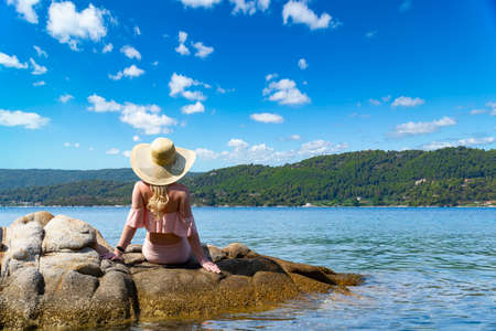 Beautiful young woman in hat sit on the cliff by the beautiful sea. Back view of sexy woman sitting by the sea in Greeceの写真素材