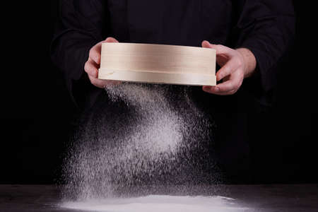 chef a man in a black uniform holds a round wooden sieve in his hands and sifts white wheat flour on...