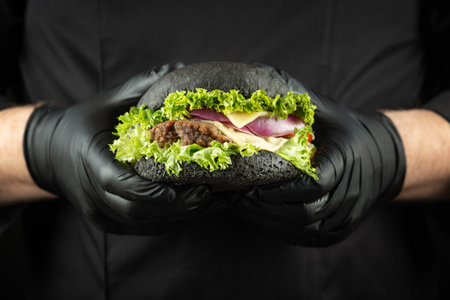 Male hands holding fresh delicious beef burger. Man in the black chef uniform serving homemade double patty burger with black angus beef, onion, tomato, cheese, lettuce, sauce on the black plate.の写真素材