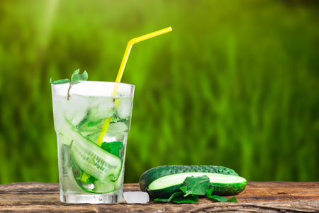 Refreshing drink with cucumber and mint, in a glass with a straw, next to ice cubes, against a green backgroundの写真素材