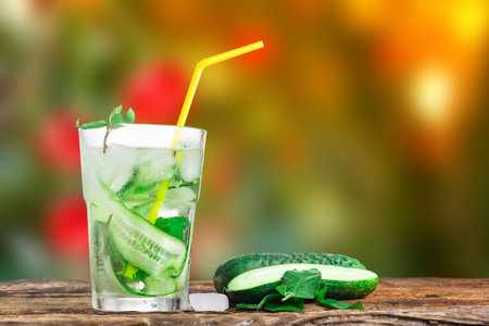 Refreshing drink with cucumber and mint, in a glass with a straw, next to ice cubes, against a green backgroundの写真素材