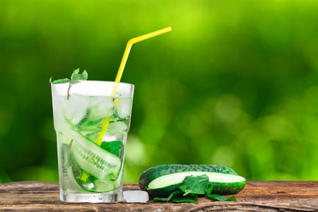 Refreshing drink with cucumber and mint, in a glass with a straw, next to ice cubes, against a green backgroundの写真素材