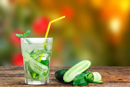 Refreshing drink with cucumber and mint, in a glass with a straw, next to ice cubes, against a green backgroundの写真素材