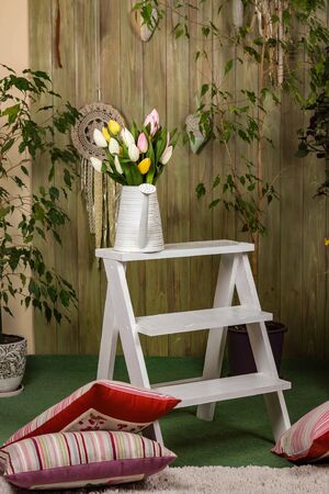 Tulips in a white watering can on the white stairs. Interior in Provence style. Home plants and green wooden wall with handmade decor.の写真素材
