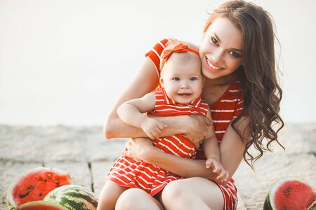 Portrait of young beautiful mother and her baby girl sitting on the beach on sunset. Woman hugging...