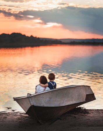 two children in the summer sit in a boat on the lake and watch a beautiful pink sunset, rear viewの写真素材