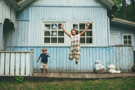 front view of boy and girl of school age joyfully jumping near their wooden blue near the forest in...