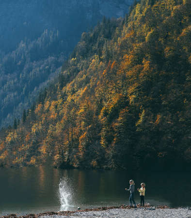 children in sunny weather throw stones at a large lake surrounded by mountains and autumn trees side viewの写真素材