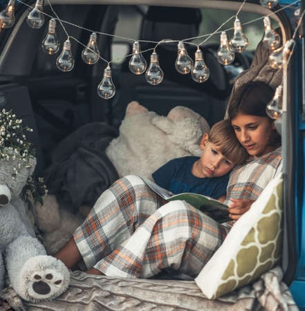 children a boy and a girl are sitting in the trunk of a car with lights, garlands with toys and a cozy blanket in the summer and reading a bookの写真素材