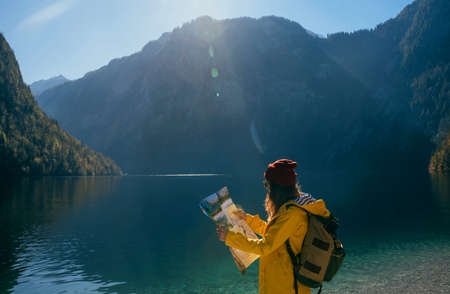 a girl in a yellow jacket and a red hat with a backpack in sunny weather looks at a map in the mountains near a blue turquoise lake side viewの写真素材