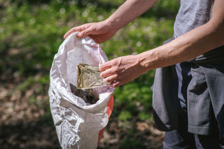 a guy in gray clothes in a green spring forest with white flowers in a sunny weather collecting trash in a bag close-upの写真素材