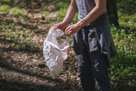 a guy in gray clothes in a green spring forest with white flowers in a sunny weather collecting trash in a bagの写真素材