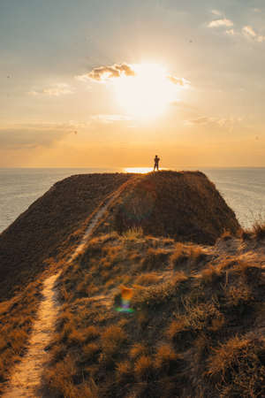a man stands on a hill with red grass on the seashore at sunset, rear viewの写真素材