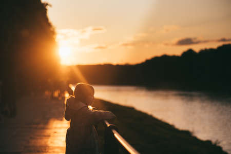 Little girl in a coat on the bridge in the sun. background of the river, trees and sunset in orange colorsの写真素材