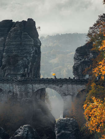 large cliffs in autumn and a stone bridge on which a man stands with a yellow umbrella front viewの写真素材