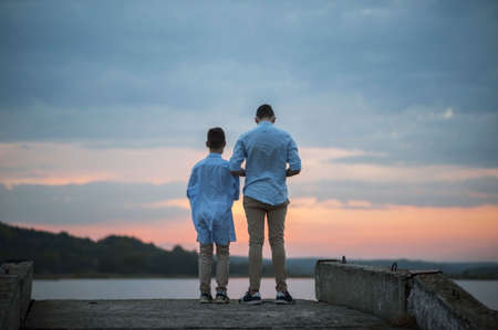 two young guys in blue shirts stand on a background of pink sunset, rear view general planの写真素材