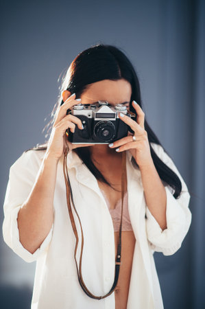 young beautiful brunette with long hair girl in a white shirt with a camera near the face takes pictures in a room against a blue wall , front view the average planの写真素材