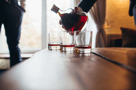 A man in a suit holds a bottle of alcohol in his hands and pours it into a glass indoors, close-up side viewの写真素材