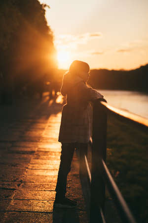 Little girl in a coat on the bridge in the sun full length. background of the river, trees and sunset in orange colors verticalの写真素材