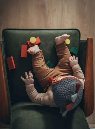 little boy in brown overalls in a knitted hat sits on a green armchair with toy wooden cubes and plays, indoors with vintage style, top view close-upの写真素材