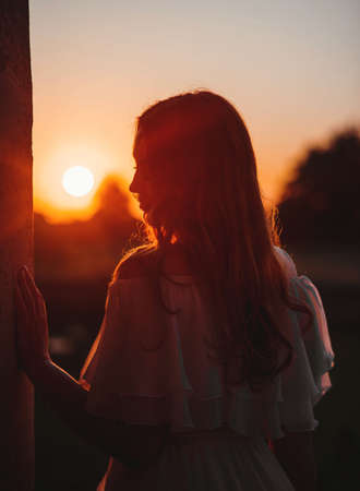 silhouette of a beautiful young girl with red curly hair in a summer dress, she stands dreamily, closing her eyes, at sunset, rear view medium planの写真素材