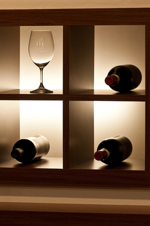A single empty wine glass with beautiful reflections and three closed bottles of red wine lying on a stand with side lighting set in the interiorの写真素材
