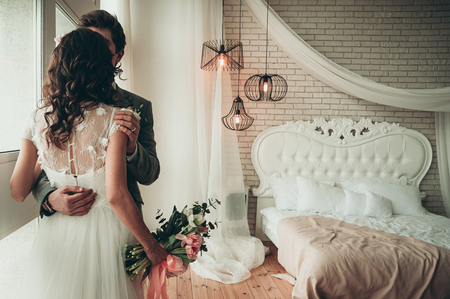 Bride and Groom with a wedding bouquet standing by the window, view from the back, a white bed in the backgroundの写真素材