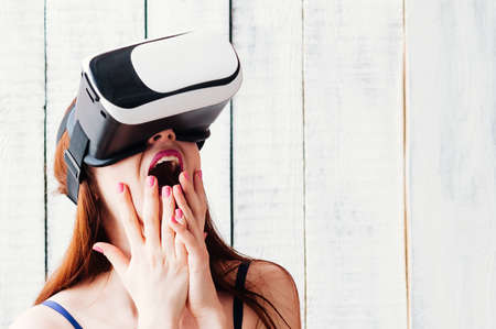 A redhead pretty young woman wearing virtual reality glasses, excitedly holding her hands at her face white wooden background, bare shoulders, pink lips and nailsの写真素材
