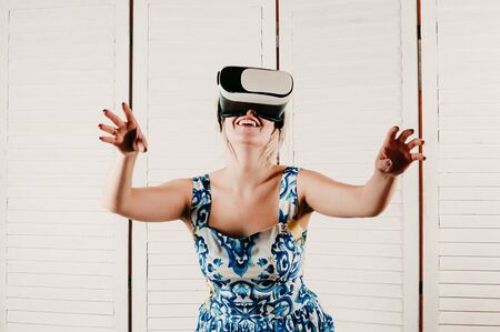 A blonde young woman wearing virtual reality glasses, smiling and pointing her hands in the air, white wooden backgroundの写真素材