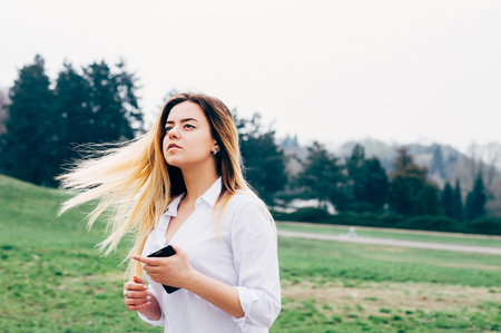 A portrait of a pretty young long haired woman in white shirt, standing in the windy park and looking into the distance, holding a cellphone in her handの写真素材