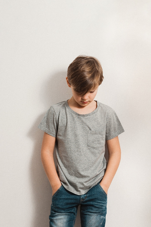 a cute boy stands next to white wall, grey t-shirt, blue jeans, hands in pockets, bowing his head downの写真素材