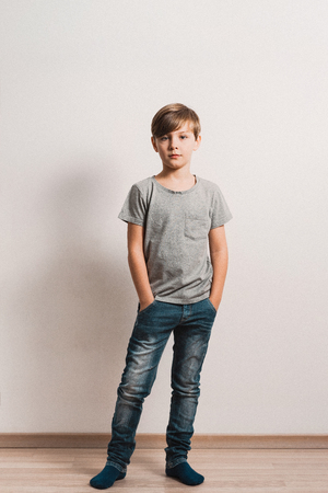 a cute boy stands next to white wall, grey t-shirt, blue jeans, hands in pockets