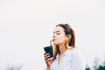 a portrait of a pretty long haired young girl wearing white shirt, drinking a coffee or smoothie through a sipping straw, smiling, eyes closed, outdoors. Copy spaceの写真素材